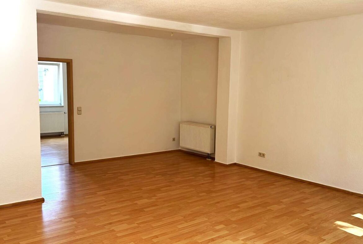 Empty living room with warm wood flooring, white walls, and a radiator on the far wall; doorway leads to a sunlit room with a window.