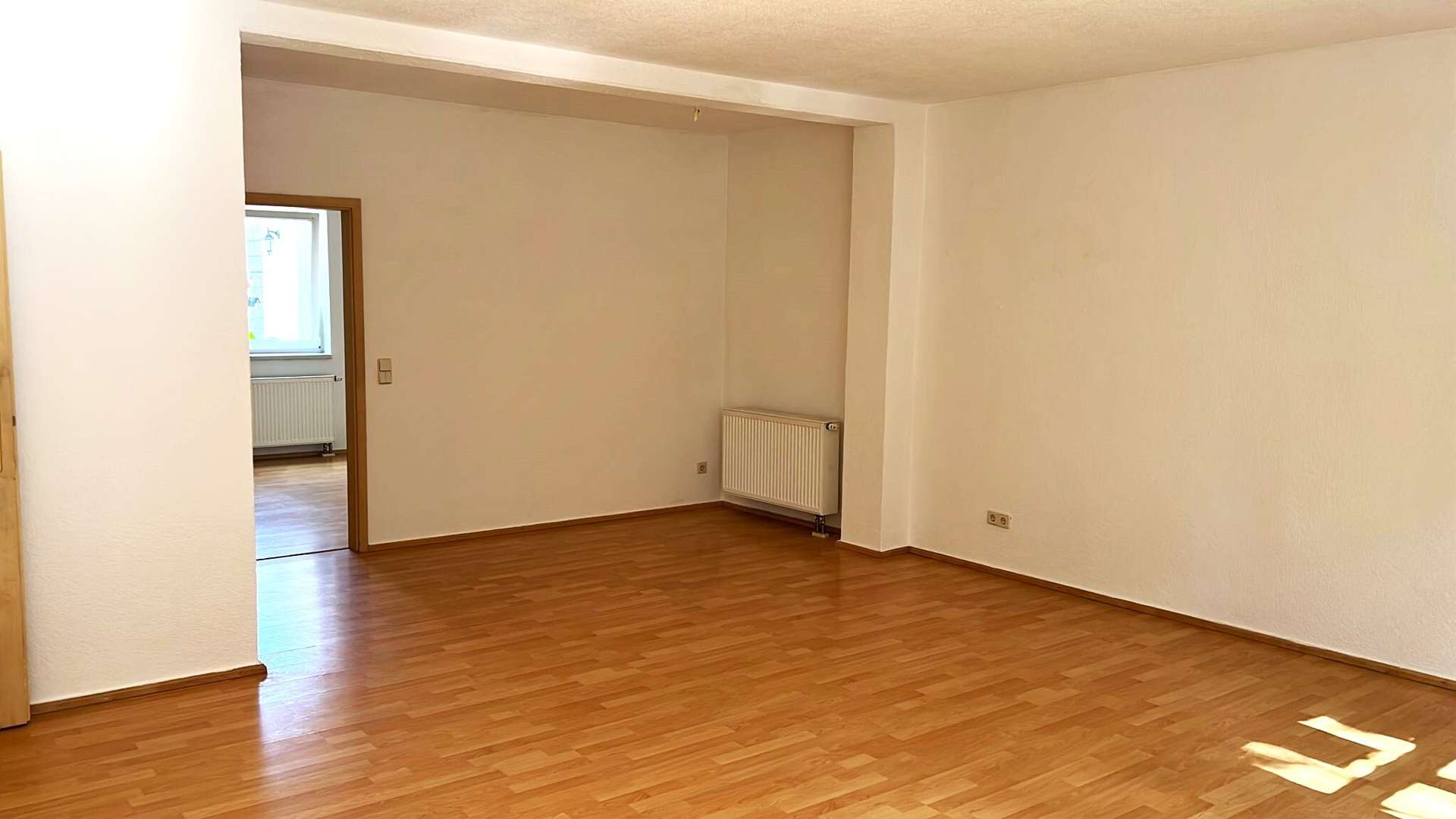 Empty living room with warm wood flooring, white walls, and a radiator on the far wall; doorway leads to a sunlit room with a window.