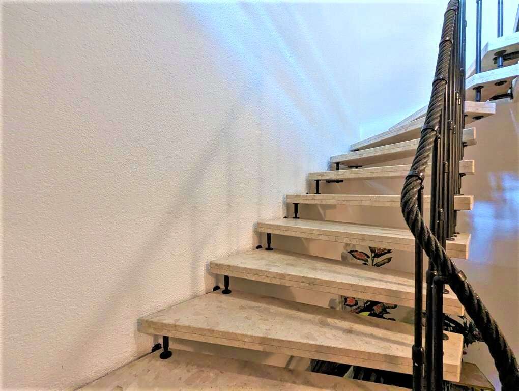 Interior staircase with light-colored stone steps and a dark twisted rope handrail along a white wall, lit by natural light from above.