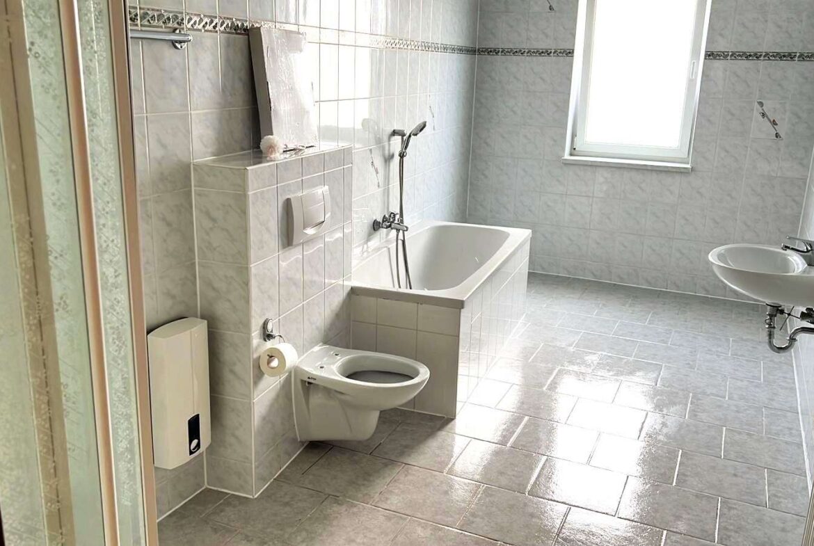 Bright gray-tiled bathroom with a white bathtub, wall-mounted toilet, and a sink by the window.