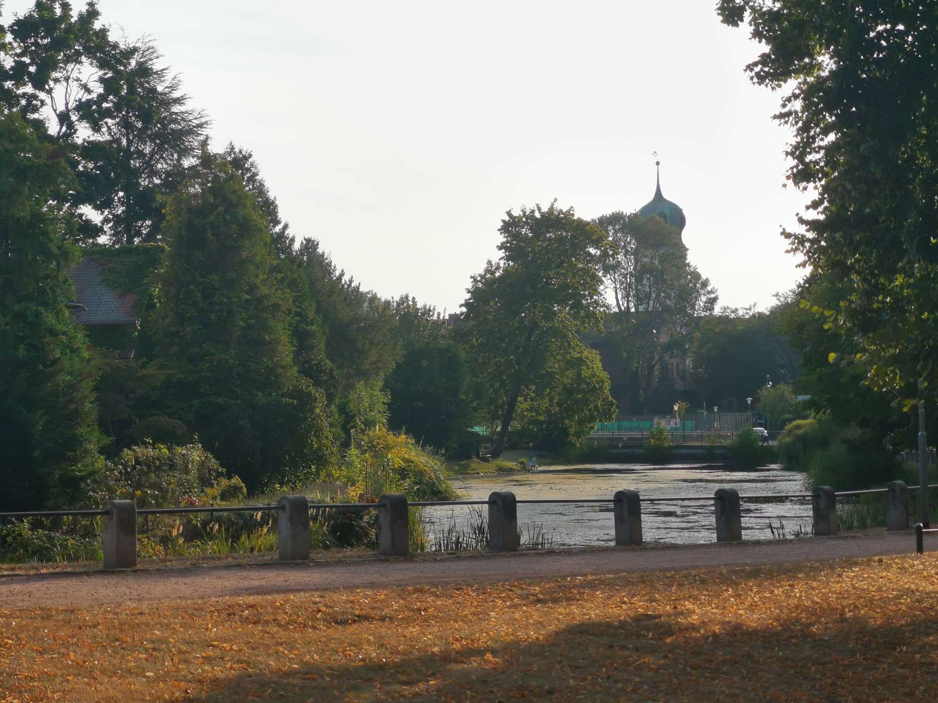 Riverbank park with a metal railing, trees, and a distant domed church tower above the treeline.