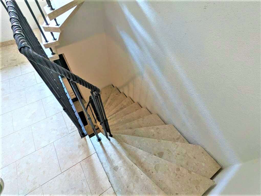 Interior stairwell seen from above: beige stone stairs with a black metal handrail along a pale wall