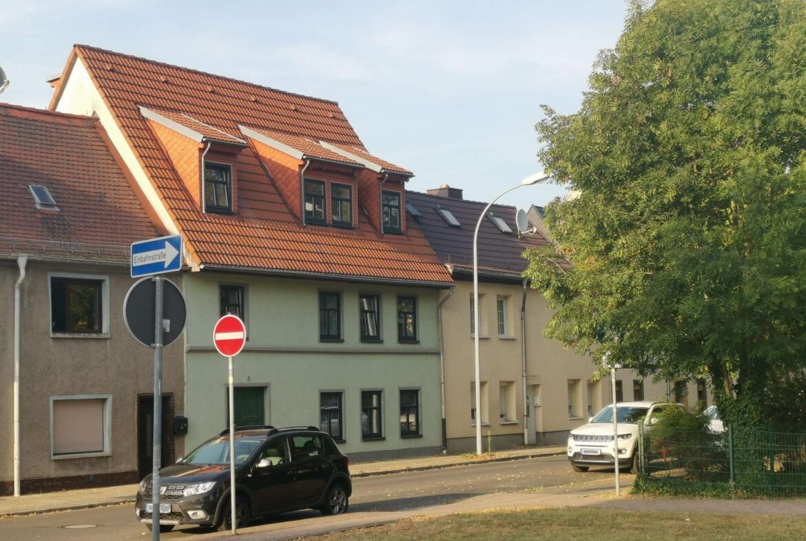 Row houses with red-tiled roofs along a street, foreground street signs including one-way and no-entry, and parked cars with a large tree on the right side of the image.