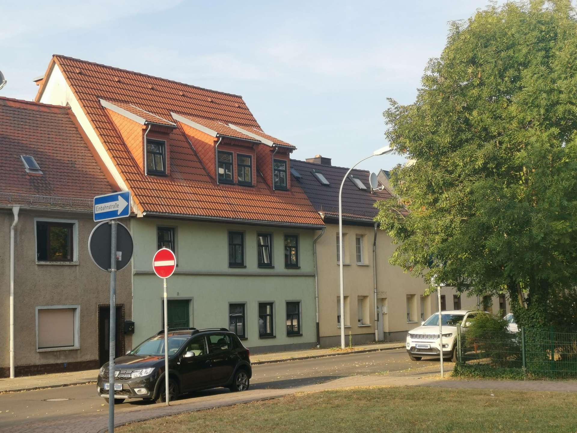 Row houses with red-tiled roofs along a street, foreground street signs including one-way and no-entry, and parked cars with a large tree on the right side of the image.