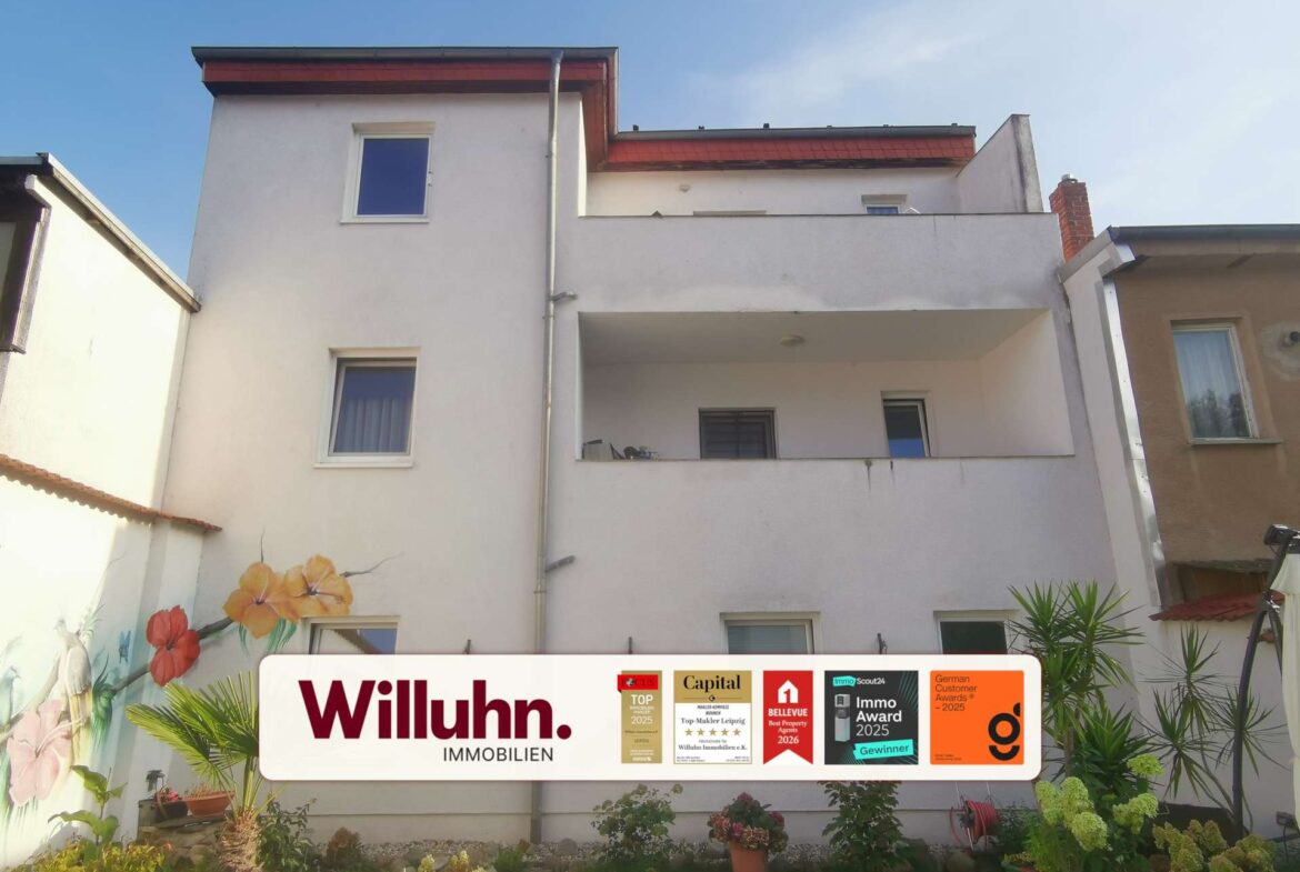 White multi-story building with a large real estate sign reading Willuhn Immobilien in a courtyard with potted plants and flowers.