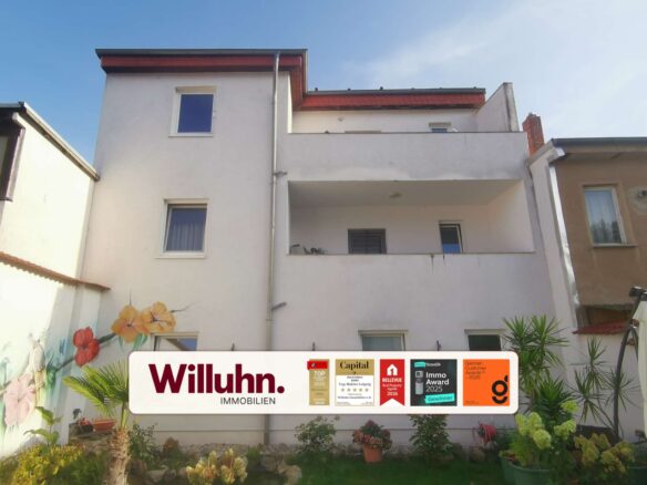 White multi-story building with a large real estate sign reading Willuhn Immobilien in a courtyard with potted plants and flowers.