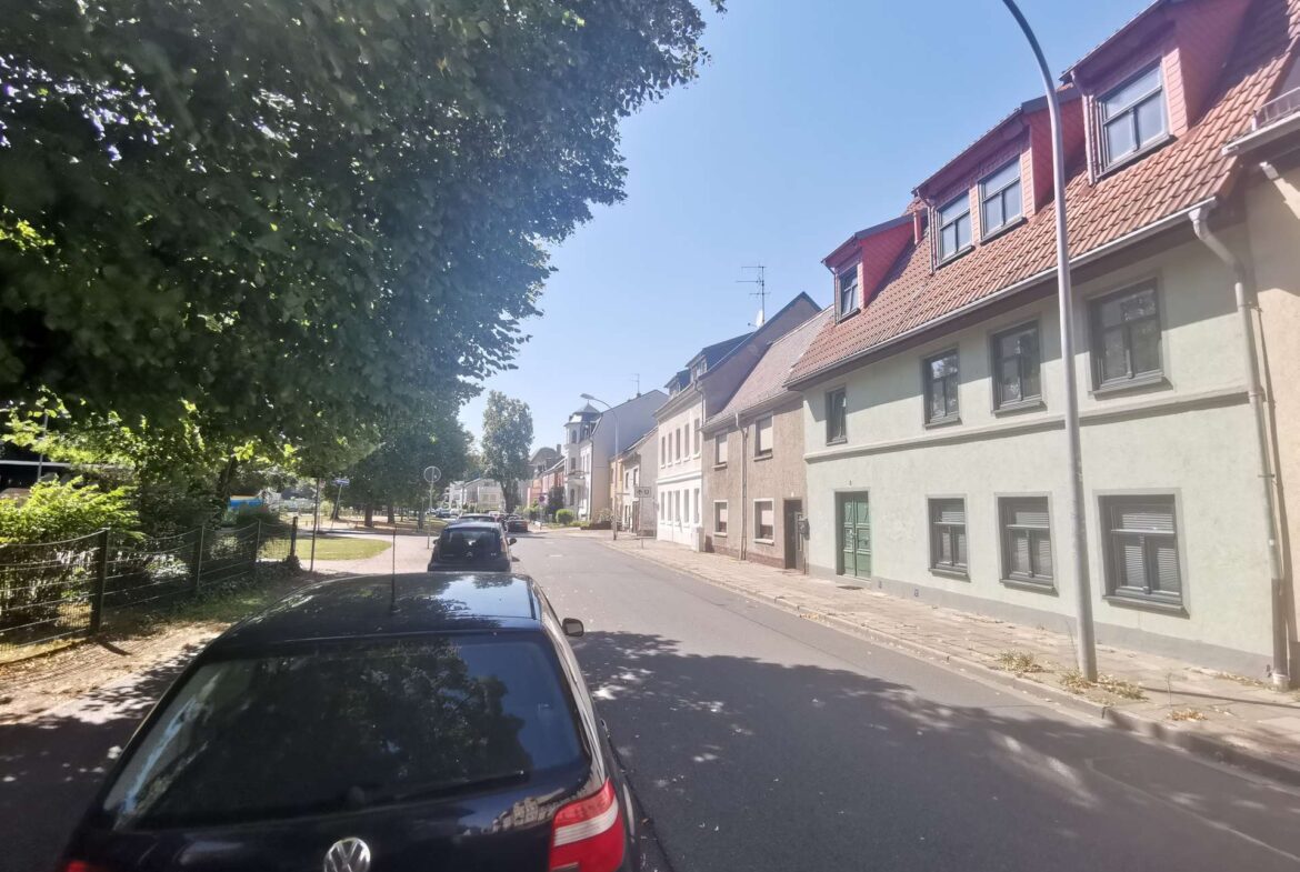 Row of beige houses with red-tiled roofs along a sunny street, parked cars on the right, and large green trees to the left under a blue sky.