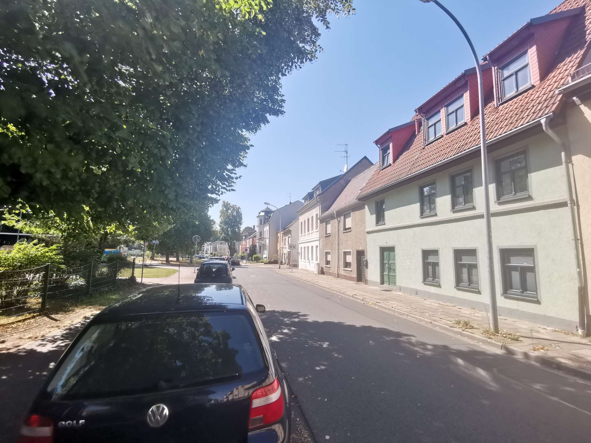 Row of beige houses with red-tiled roofs along a sunny street, parked cars on the right, and large green trees to the left under a blue sky.