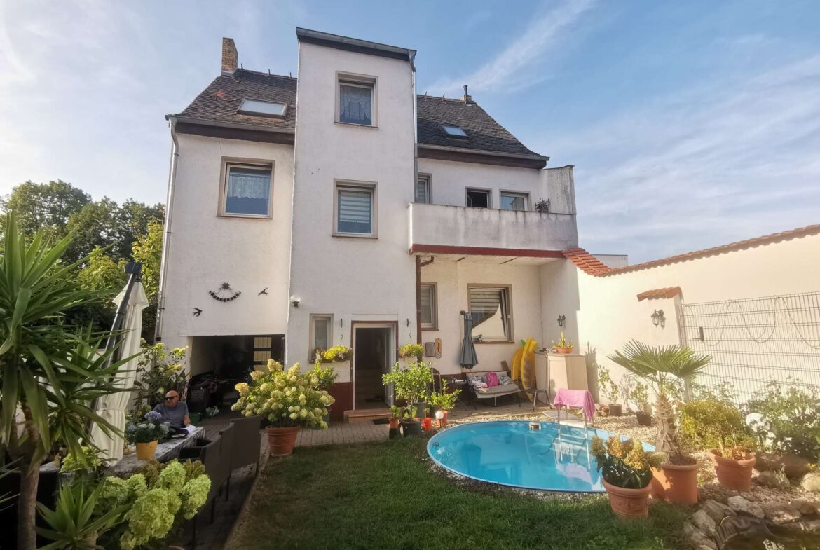 White three-story house with a terrace, surrounded by potted plants and a round blue pool in the backyard under a blue sky.
