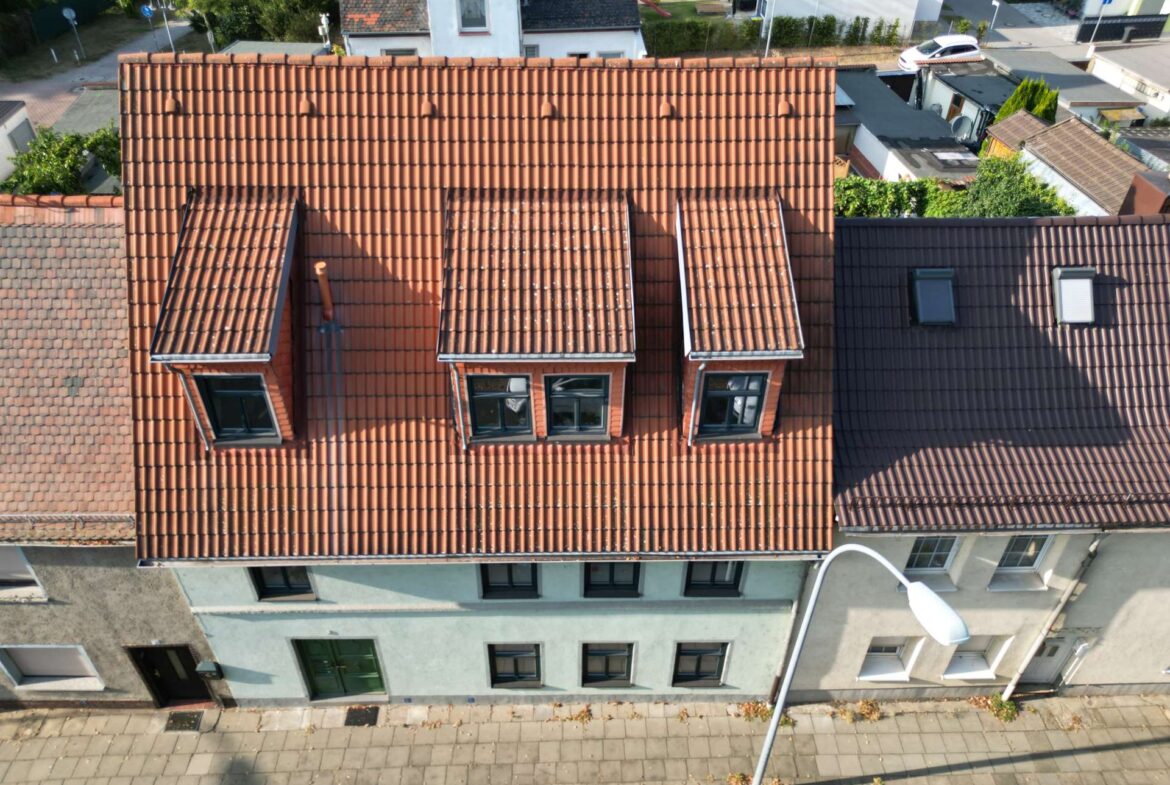 Aerial view of a red-tiled roof with three dormer windows, overlooking a street with a sidewalk and lamp post in front.