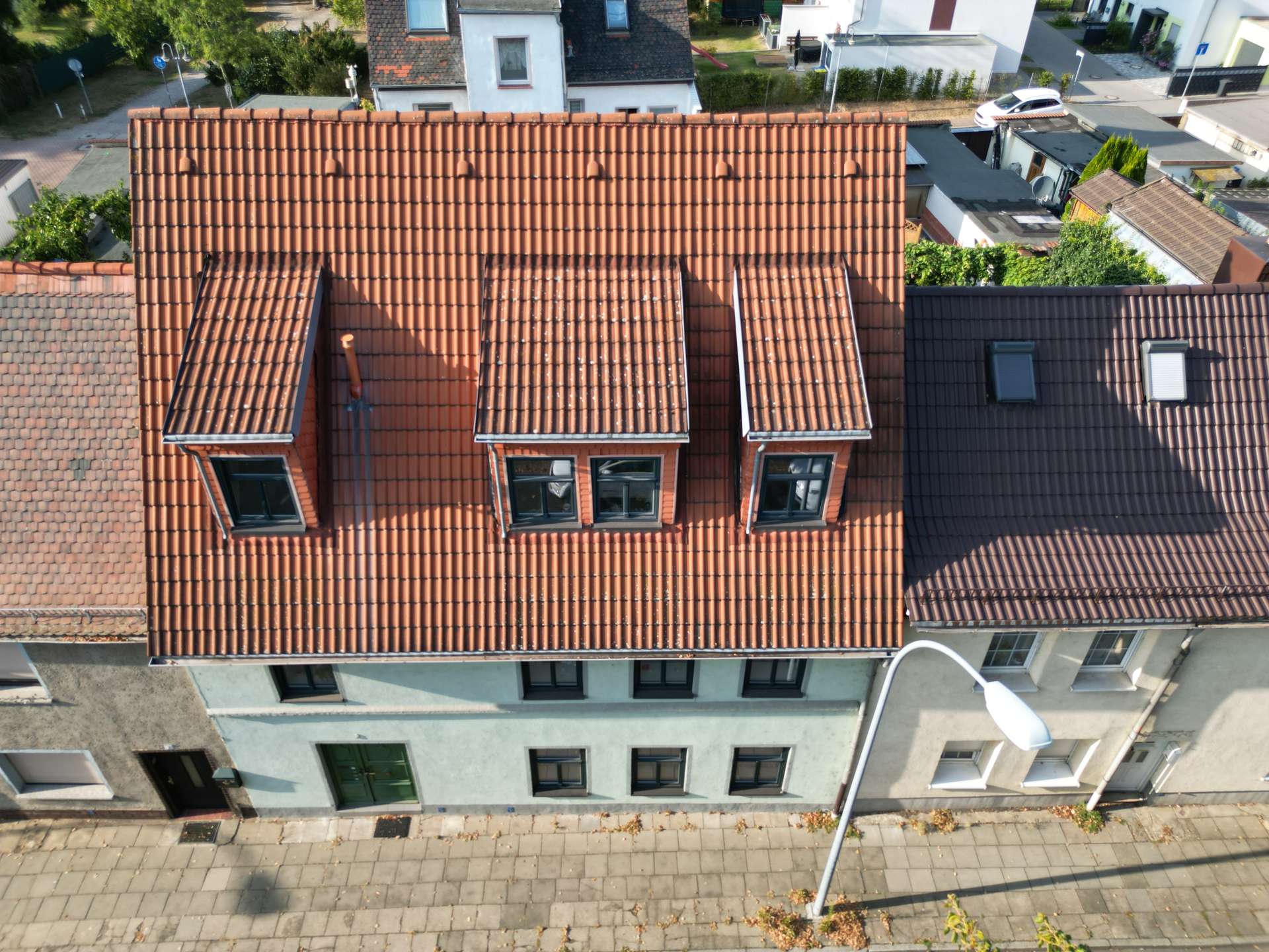 Aerial view of a red-tiled roof with three dormer windows, overlooking a street with a sidewalk and lamp post in front.