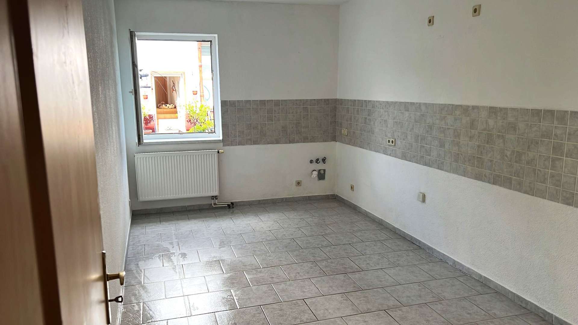 Empty kitchen with gray tiled floor and mid-wall backsplash, radiator under a window, and exposed plumbing connections on the wall center-right