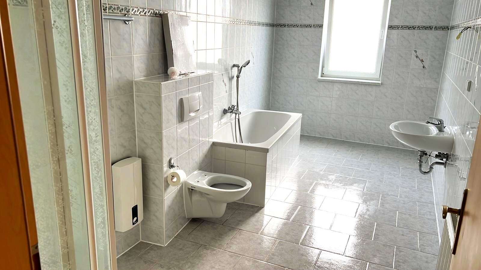 Bright tiled bathroom with a white bathtub, wall-mounted toilet, sink, and a window at the far end