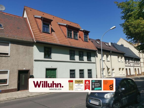 Residential street with red-tiled roofs and a large Willuhn Immobilien advertising sign in the foreground; awards stickers are visible on the sign, a car parked on the right.