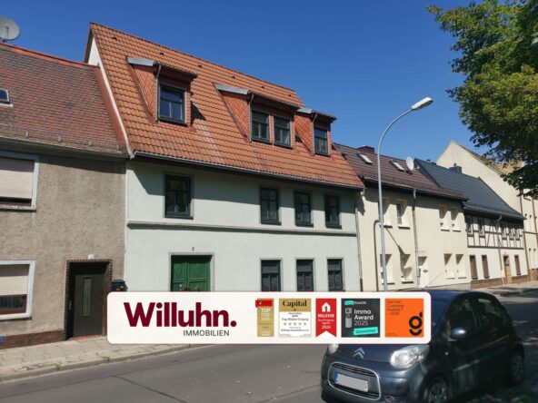 Residential street with red-tiled roofs and a large Willuhn Immobilien advertising sign in the foreground; awards stickers are visible on the sign, a car parked on the right.