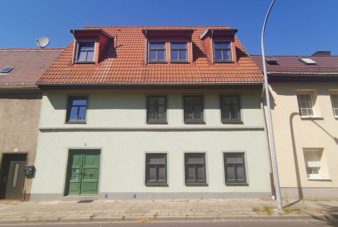 Three-story townhouse with a red tiled roof featuring dormer windows and a green front door, along a paved sidewalk.