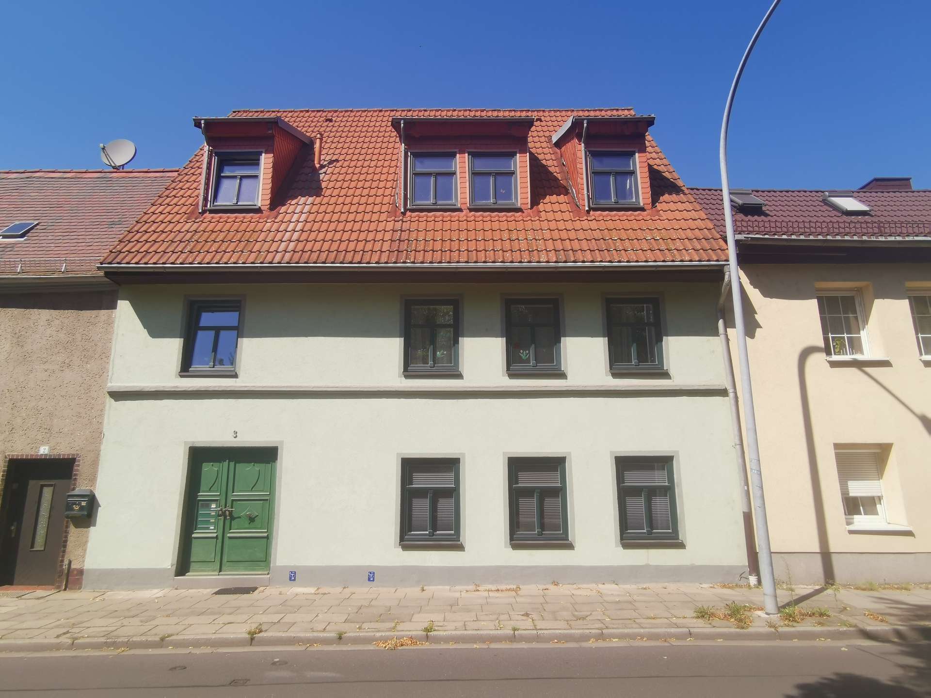 Three-story townhouse with a red tiled roof featuring dormer windows and a green front door, along a paved sidewalk.