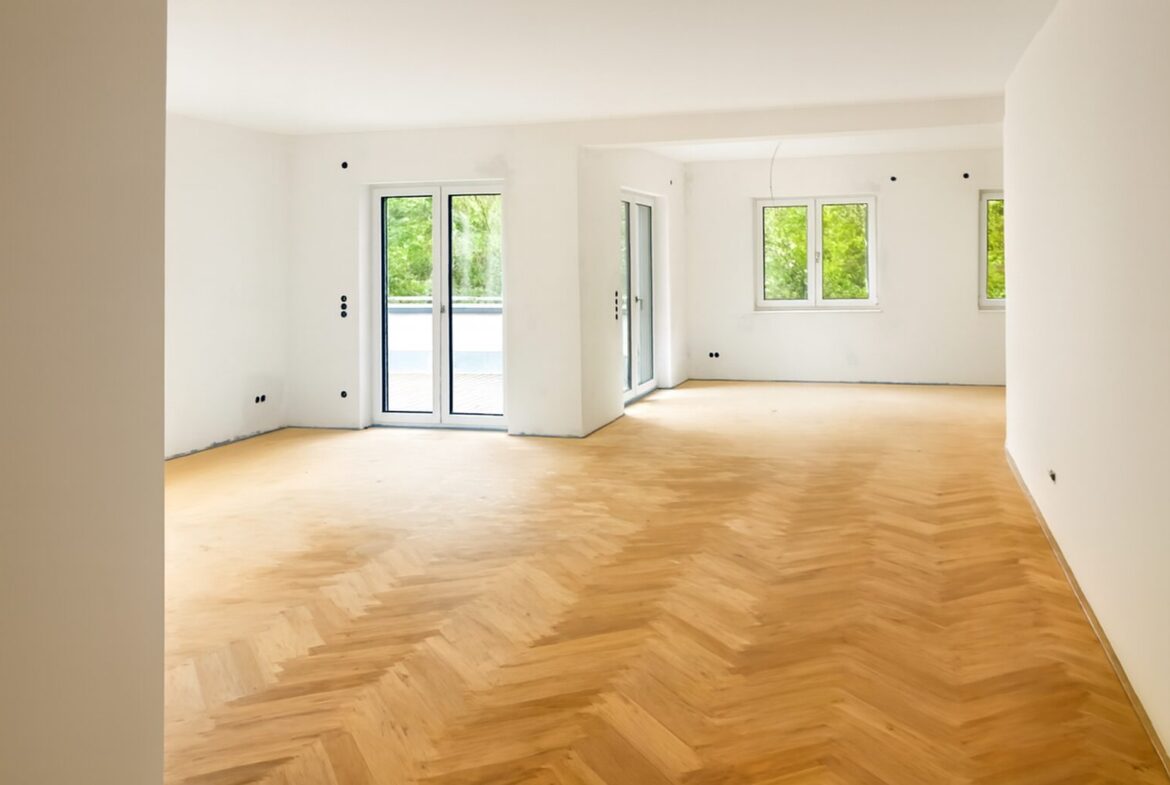 Empty white living room with a herringbone wood floor and large glass doors opening to a balcony, with greenery outside.