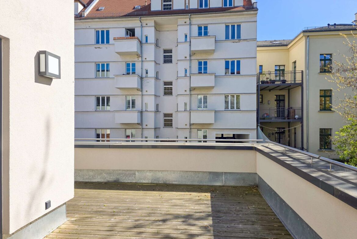 Wooden rooftop terrace with low walls and metal railing, overlooking a white multi-story apartment building with small balconies on a sunny day.