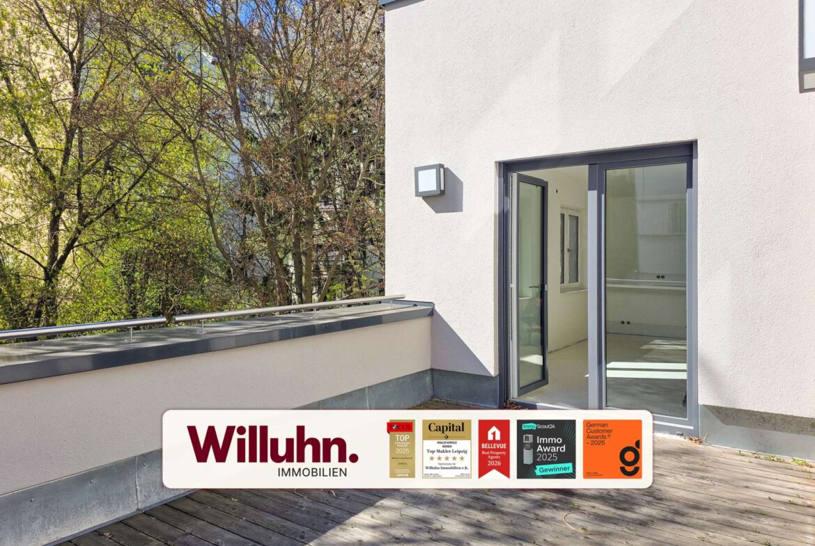 Modern white building exterior with large glass doors opening onto a wooden deck; a Willuhn Immobilien signboard with award logos rests on the deck edge.