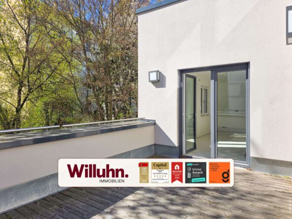 Modern white building exterior with large glass doors opening onto a wooden deck; a Willuhn Immobilien signboard with award logos rests on the deck edge.