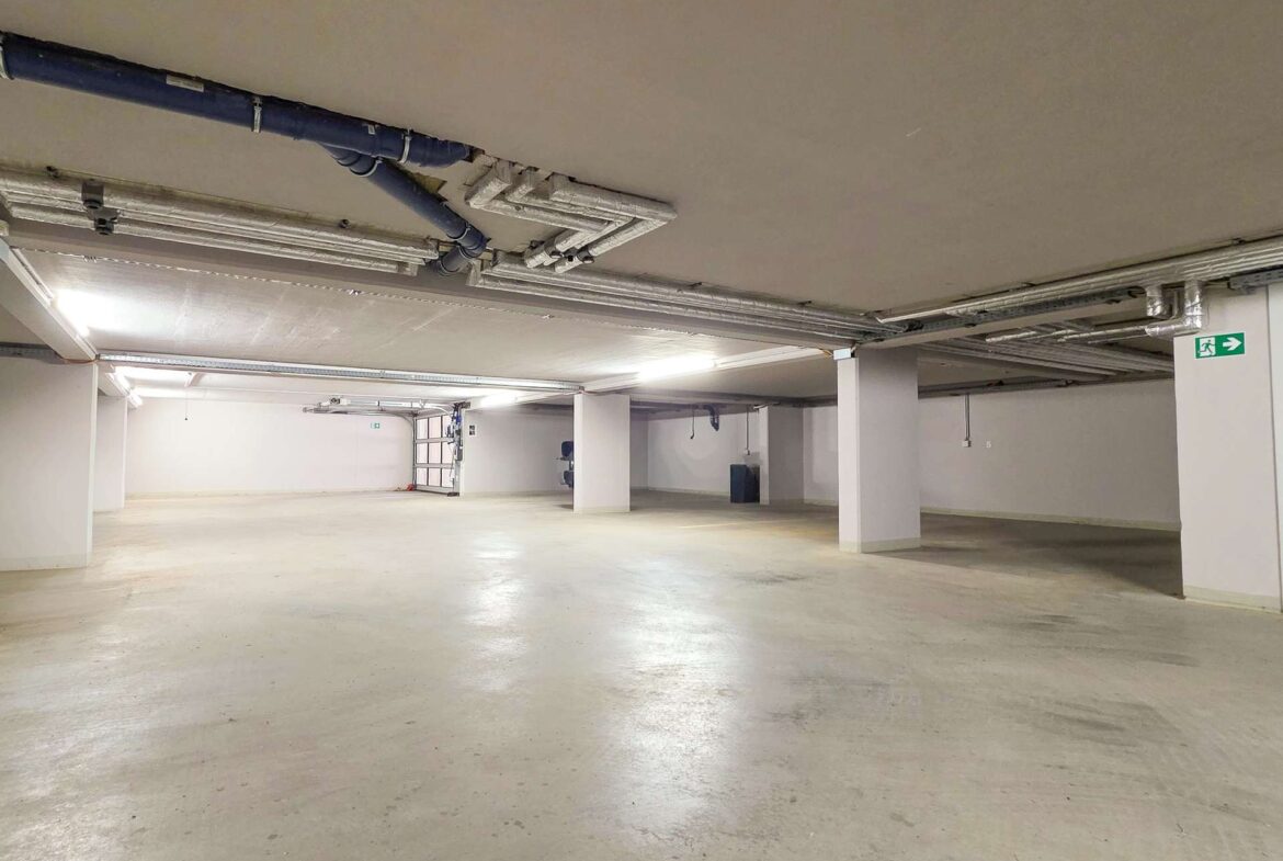 Underground parking garage with a wide empty concrete floor, exposed pipes and ductwork on the ceiling, and pillars supporting the space.