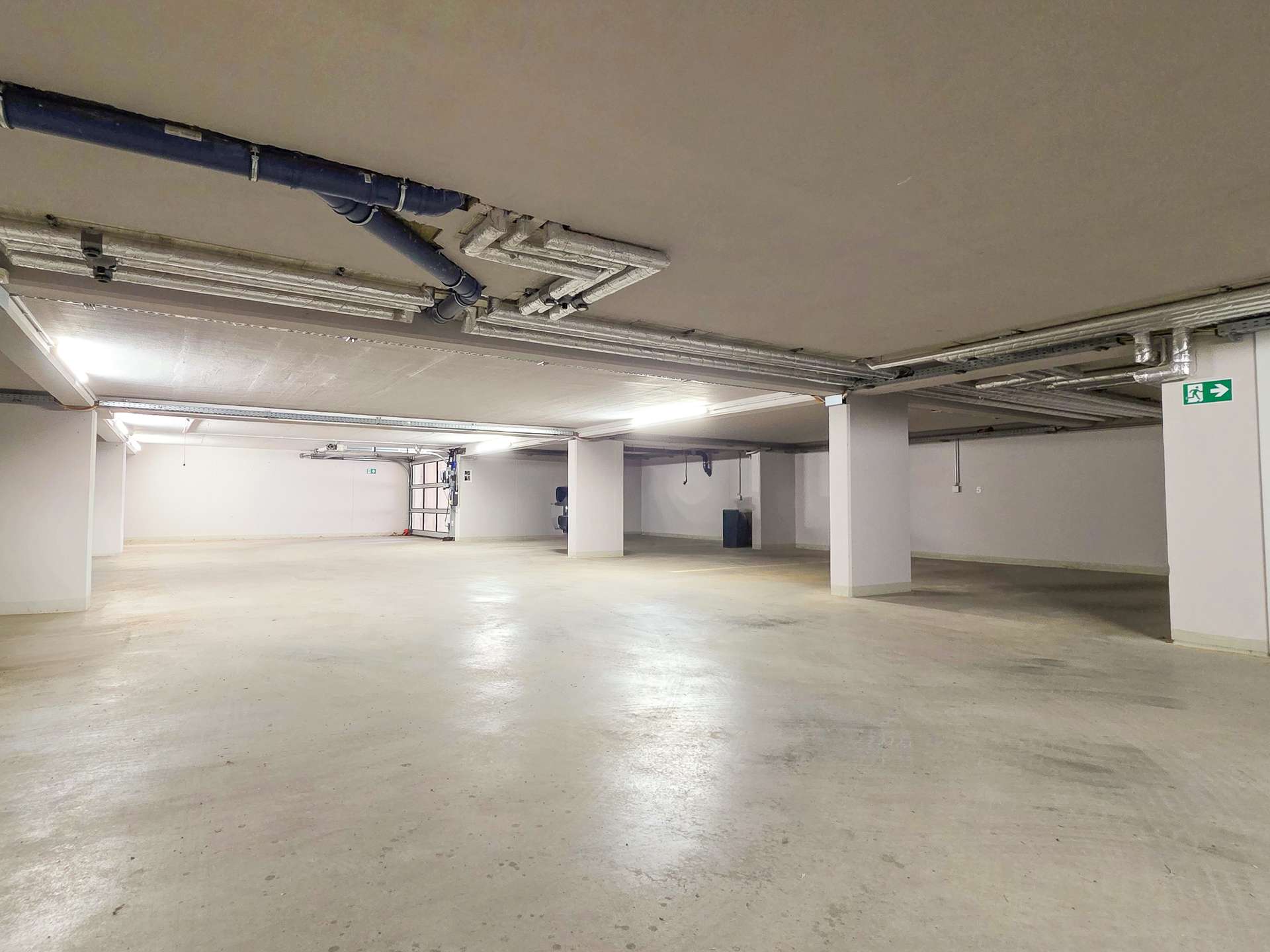 Underground parking garage with a wide empty concrete floor, exposed pipes and ductwork on the ceiling, and pillars supporting the space.