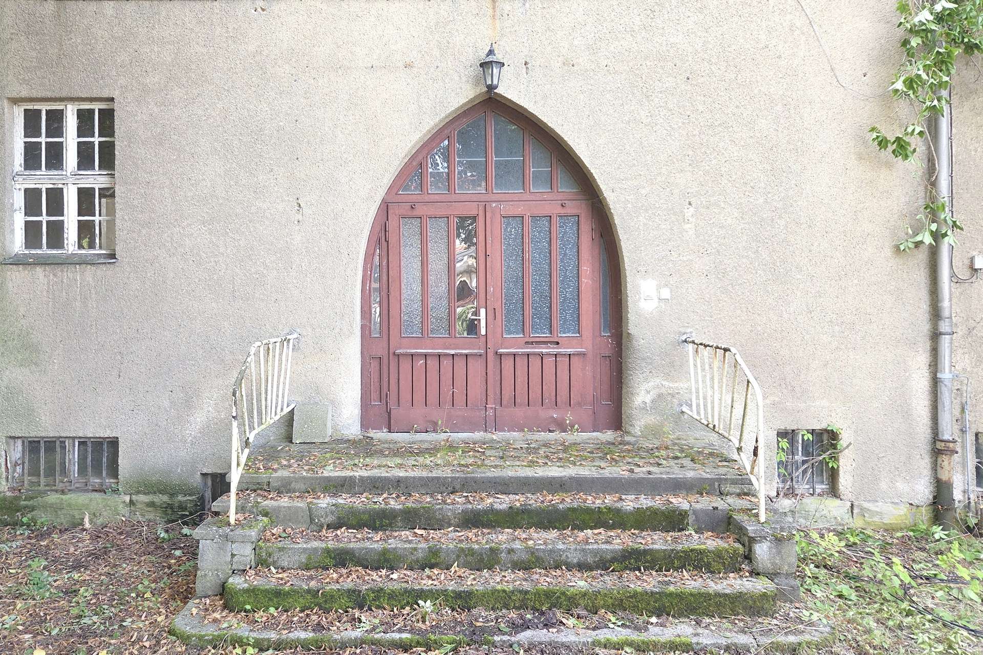 Old beige wall with a red arched double door, frosted glass panels, and a lantern above, at the top of mossy concrete steps.