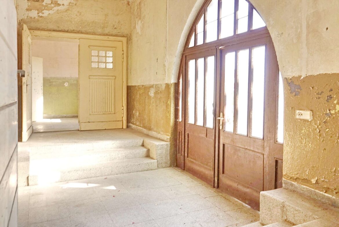 Dusty, sunlit hallway in an old building with peeling walls and a large arched glass door on the right.