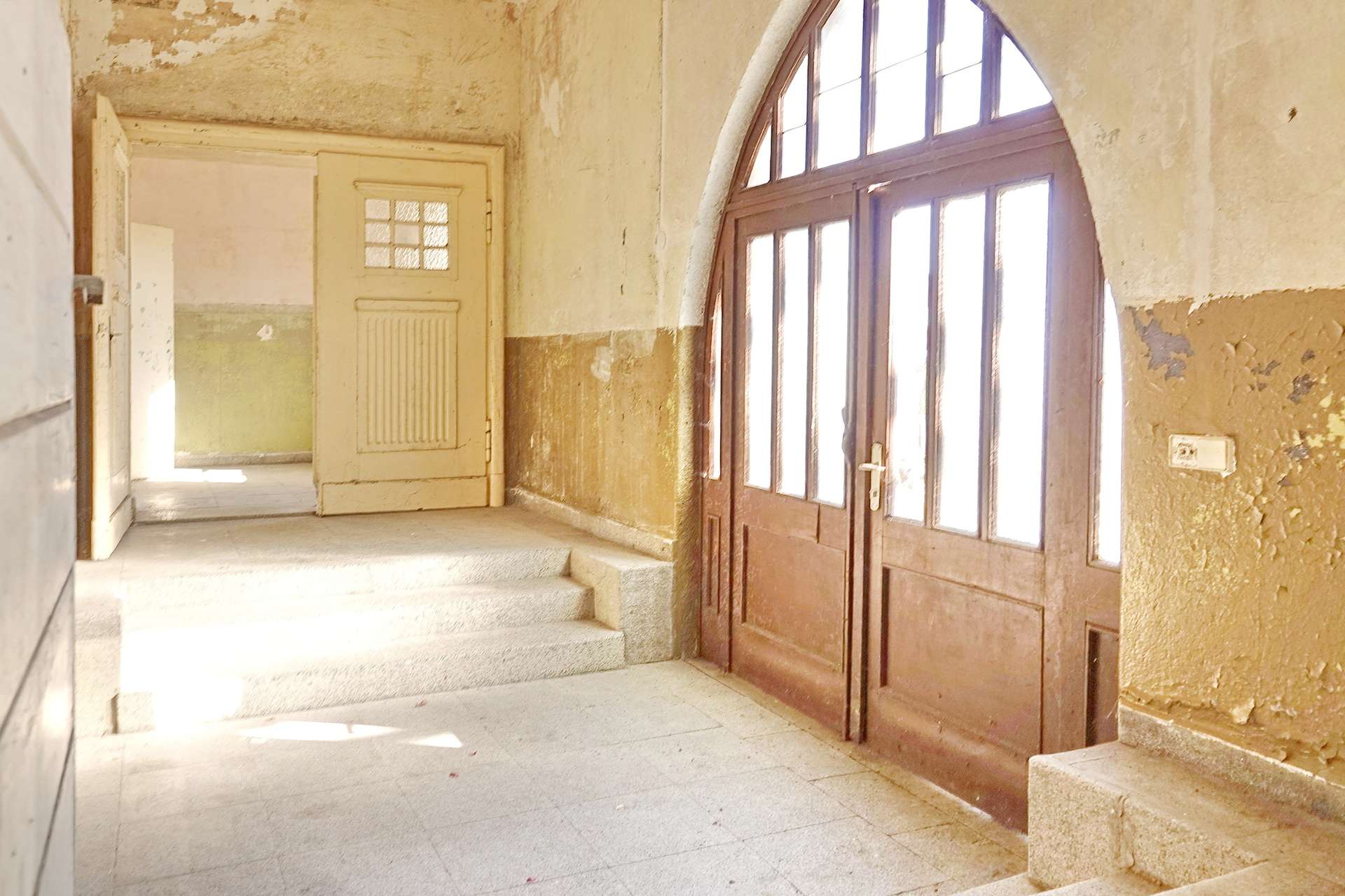 Dusty, sunlit hallway in an old building with peeling walls and a large arched glass door on the right.