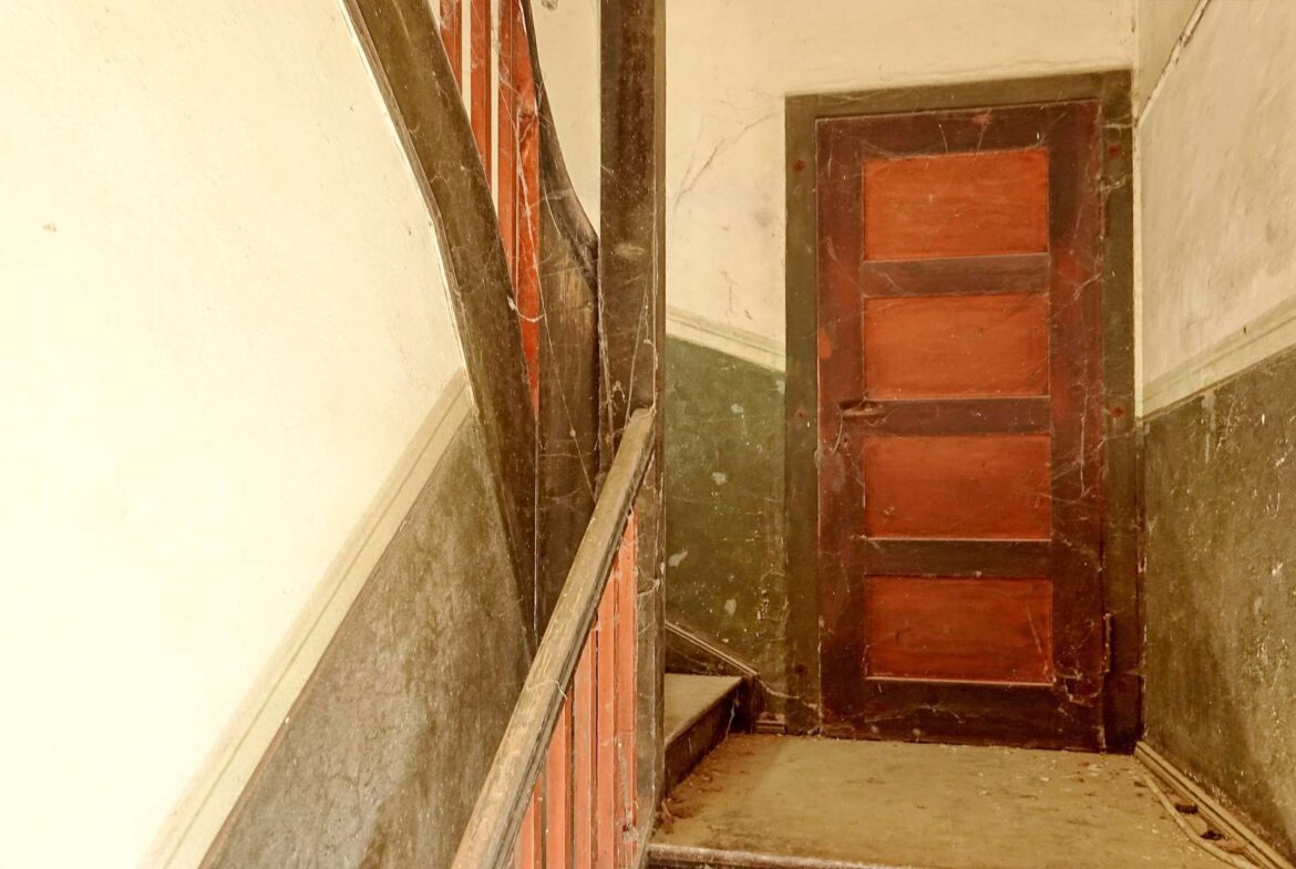 Old, dusty wooden staircase in a narrow, dim stairwell with red-brown steps and a dark handrail on the left.
