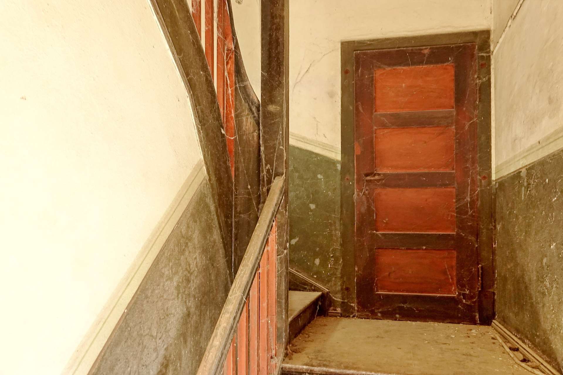 Old, dusty wooden staircase in a narrow, dim stairwell with red-brown steps and a dark handrail on the left.