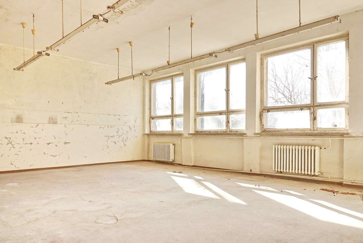 A dusty, empty industrial room with large windows letting in sunlight; peeling paint on walls and ceiling, and hanging light fixtures above a bare concrete floor.