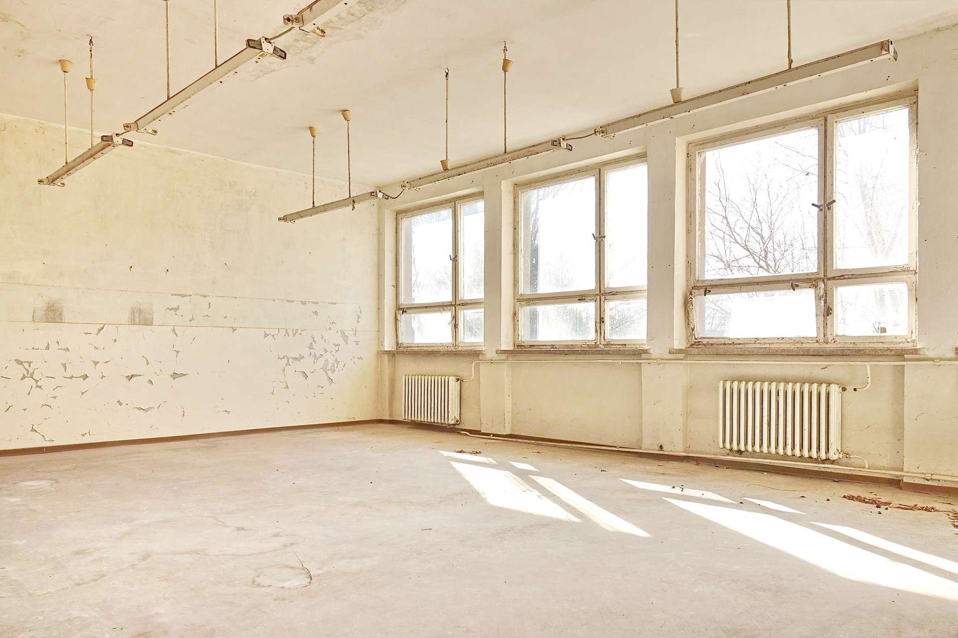 A dusty, empty industrial room with large windows letting in sunlight; peeling paint on walls and ceiling, and hanging light fixtures above a bare concrete floor.