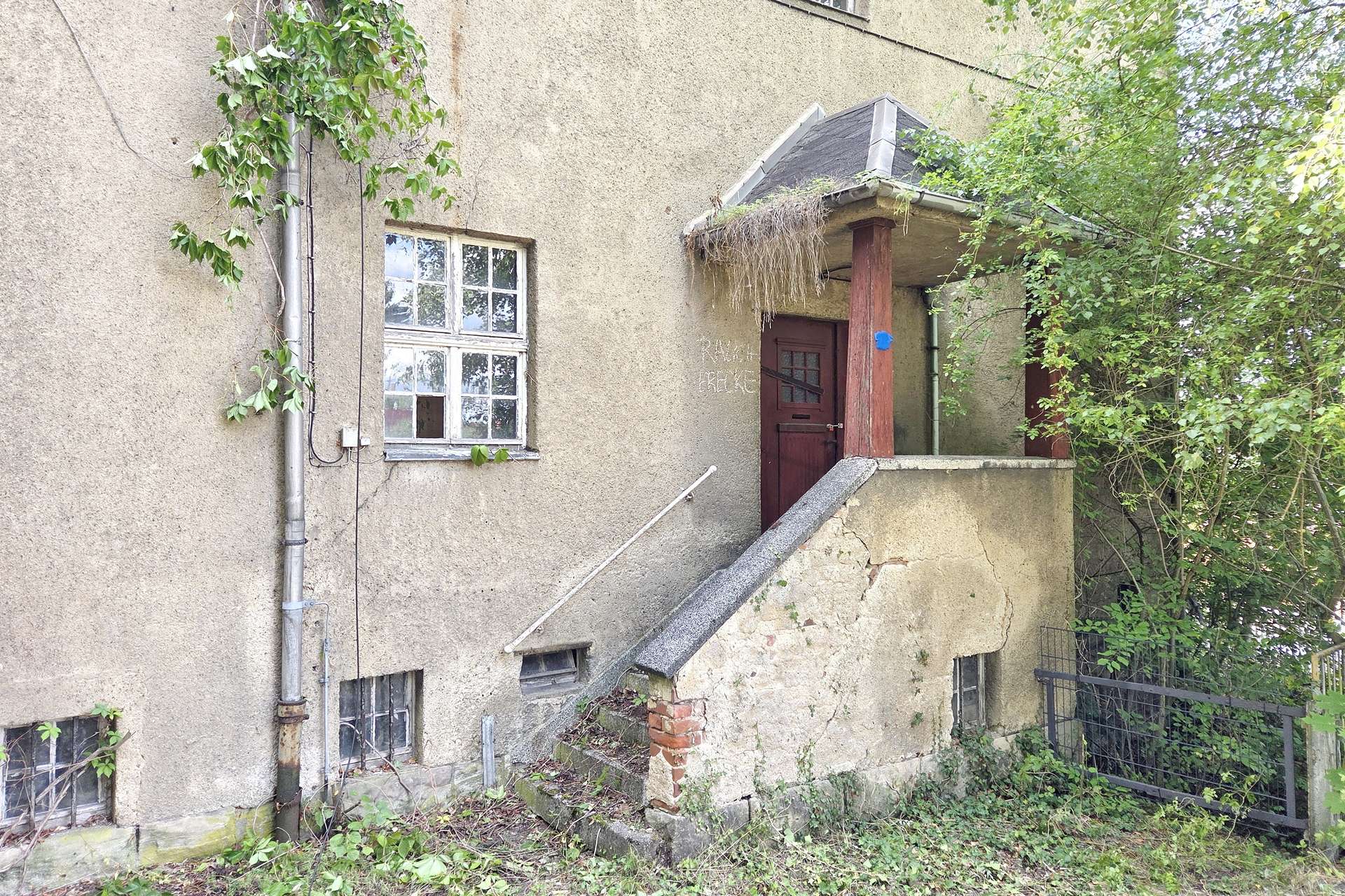 Older, beige concrete building with cracked walls, ivy growing along a metal downspout, and a stairway leading to a weathered red door under a small porch with overhanging vegetation.