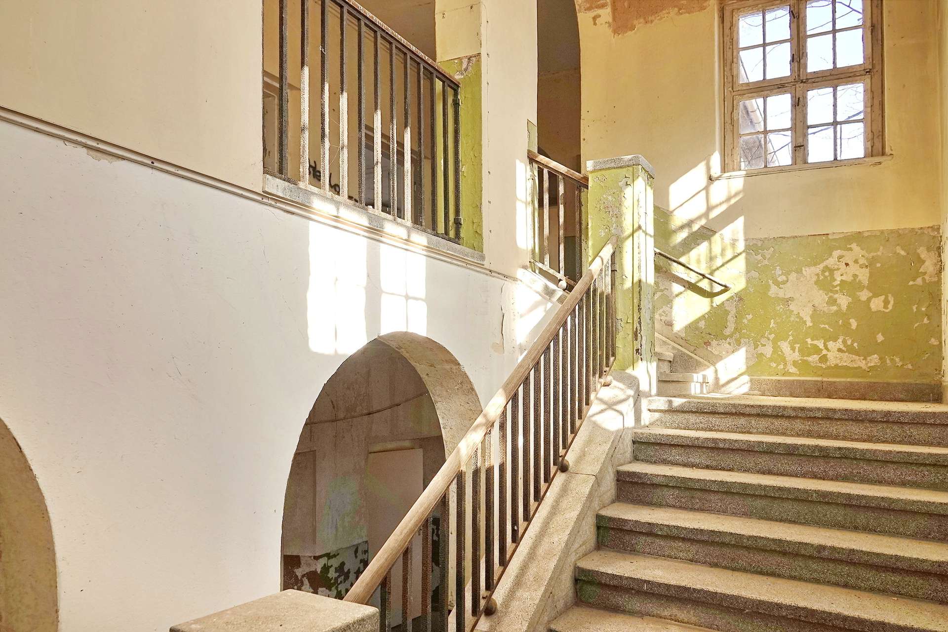 Sunlit, abandoned stairwell with a metal railing and peeling yellow walls in a worn building interior.