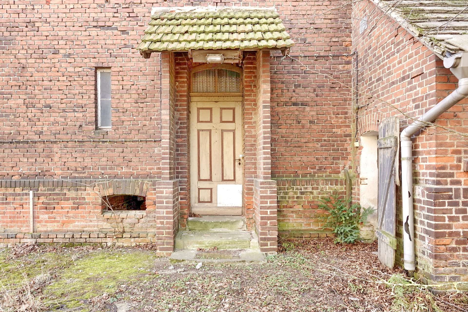 Old brick building with a wooden paneled door under a mossy tiled porch roof; overgrown yard.](informative)