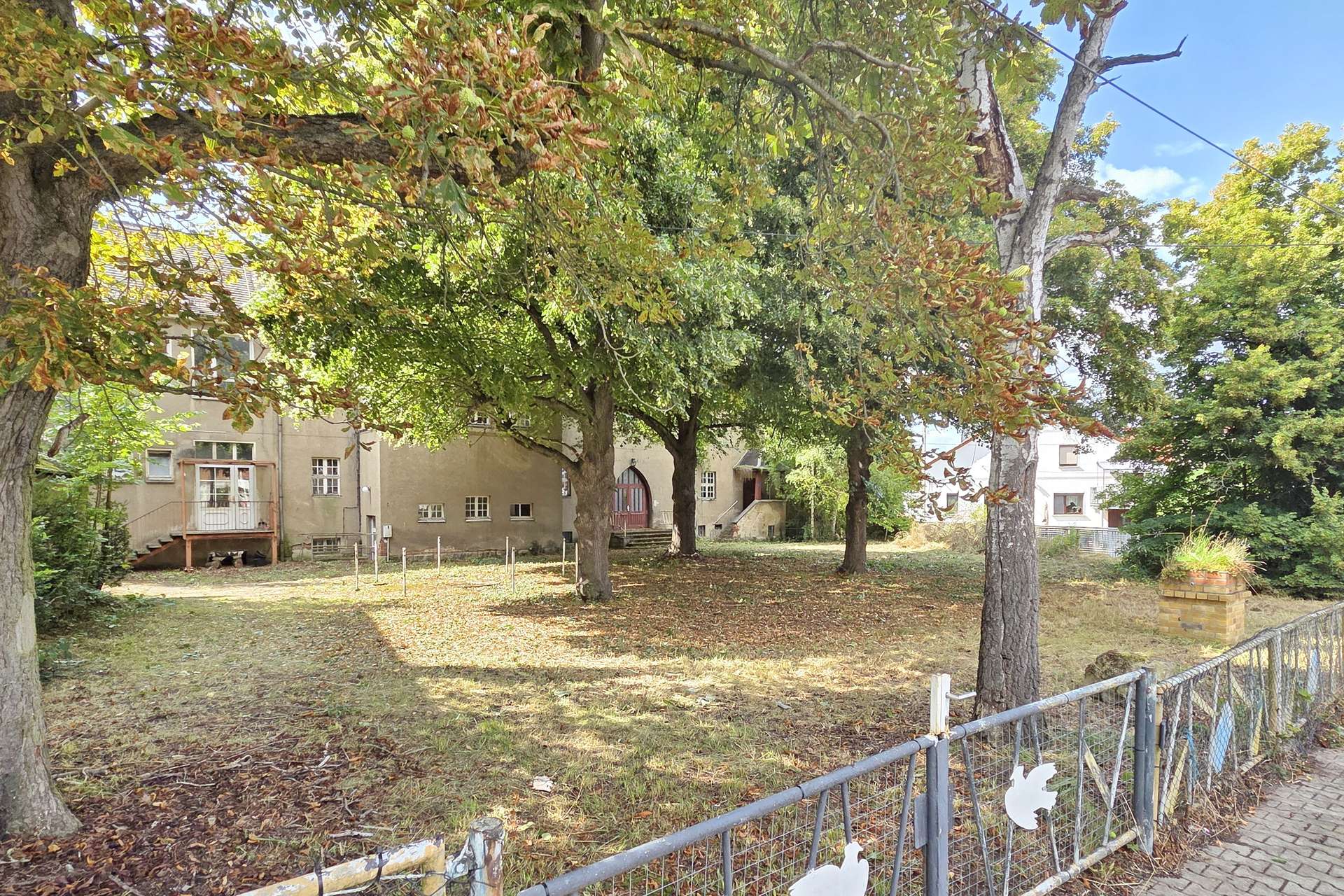 Tree-dotted yard in front of beige houses, with a metal fence and fallen leaves on the ground.