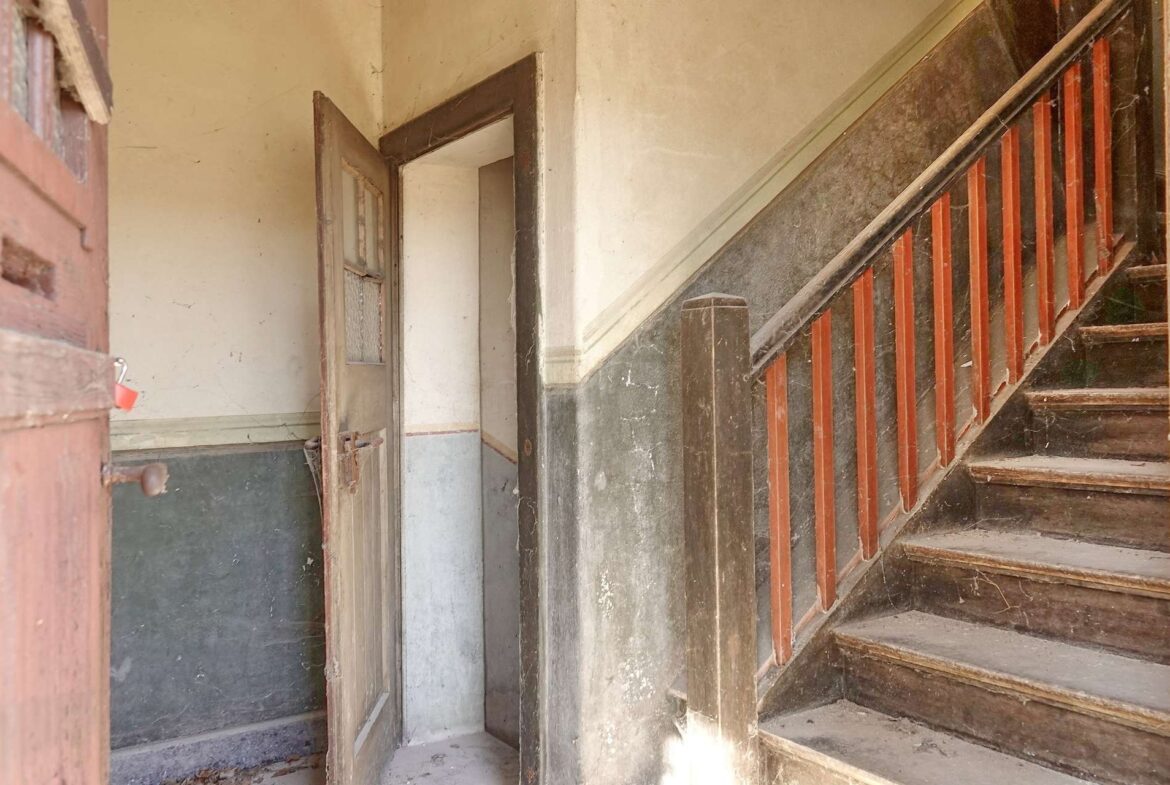Dusty, abandoned stairwell with a red wooden railing and worn steps with grime on the walls.