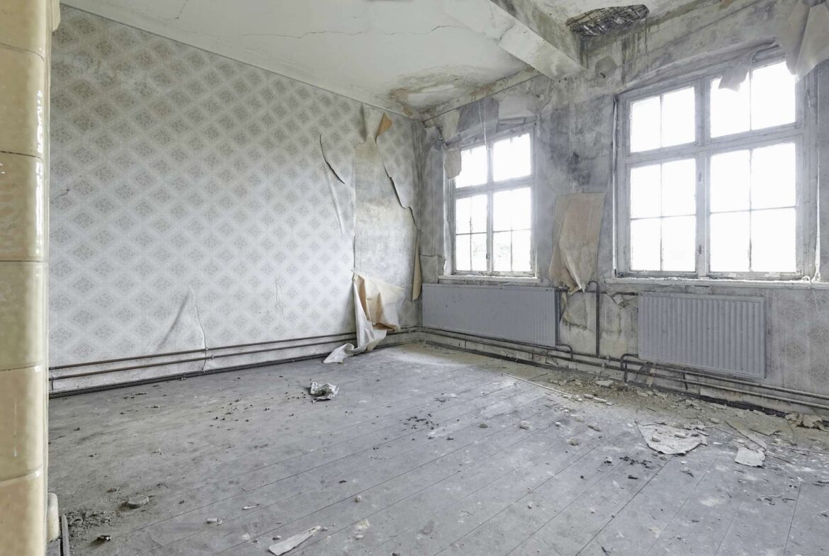 Abandoned room with peeling wallpaper, cracked walls, and debris on a dusty wooden floor near large windows to the right.