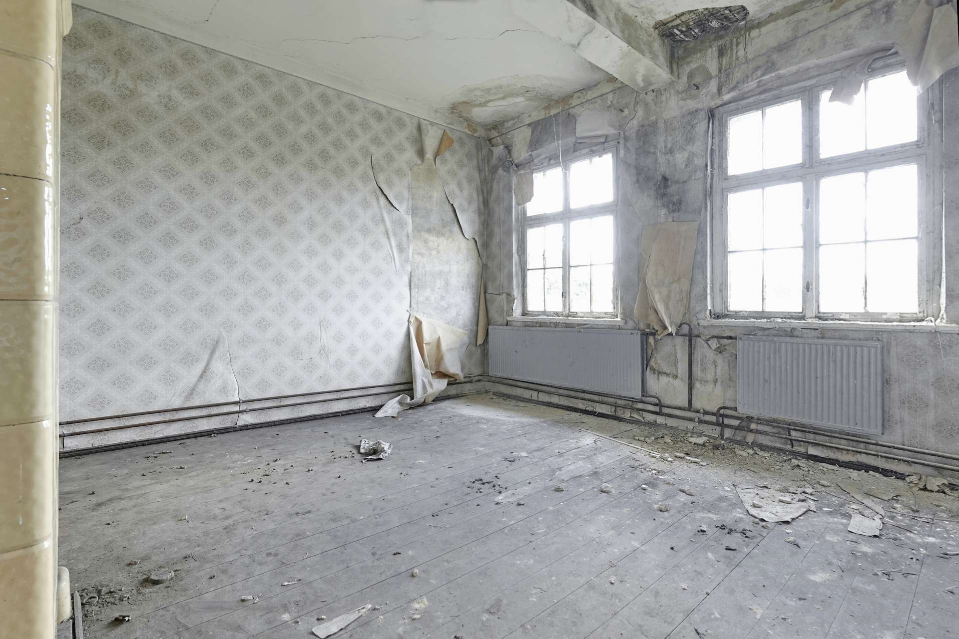 Abandoned room with peeling wallpaper, cracked walls, and debris on a dusty wooden floor near large windows to the right.