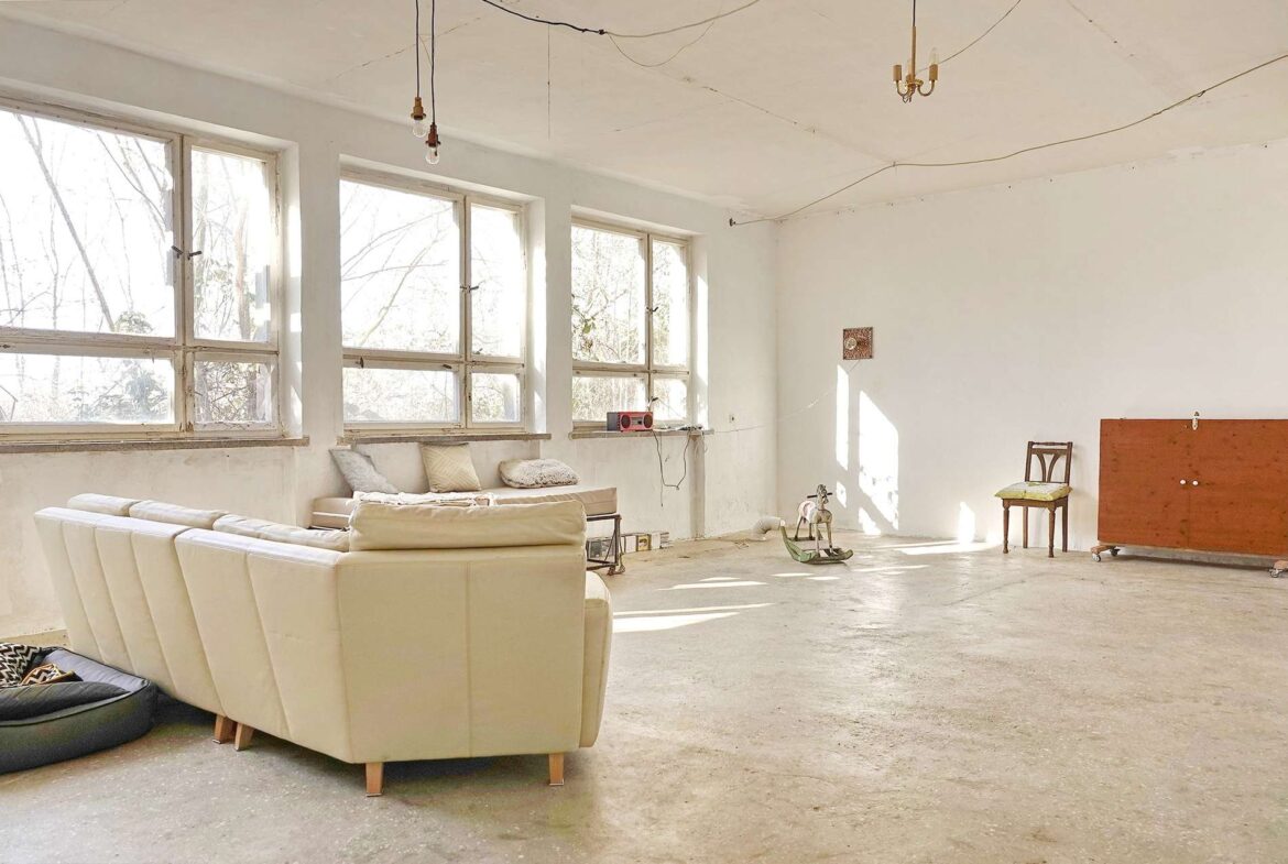 Sunlit unfinished living room with a beige sofa, cushions, and hanging light bulbs in an industrial, renovation-like space.
