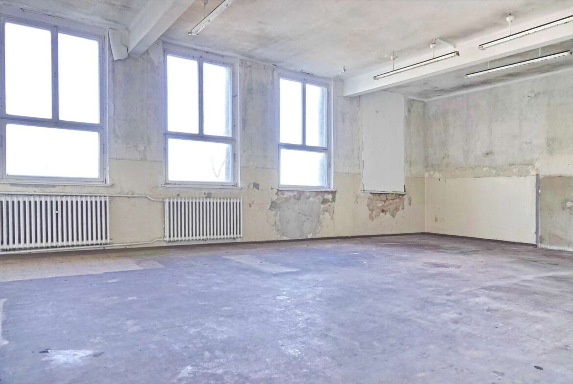 Empty industrial room with tall windows, peeling walls, and two radiators along the baseboard.