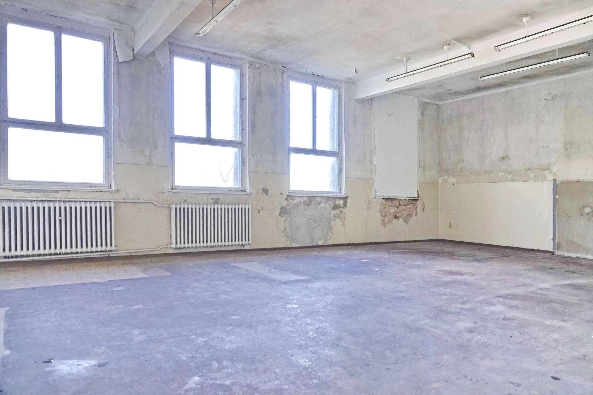 Empty industrial room with tall windows, peeling walls, and two radiators along the baseboard.