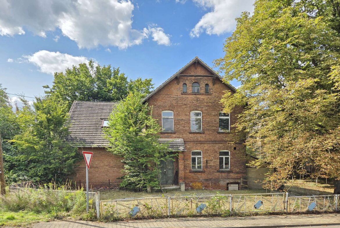 Old red brick house with a steep gabled roof and arched attic windows, flanked by green trees under a blue sky.