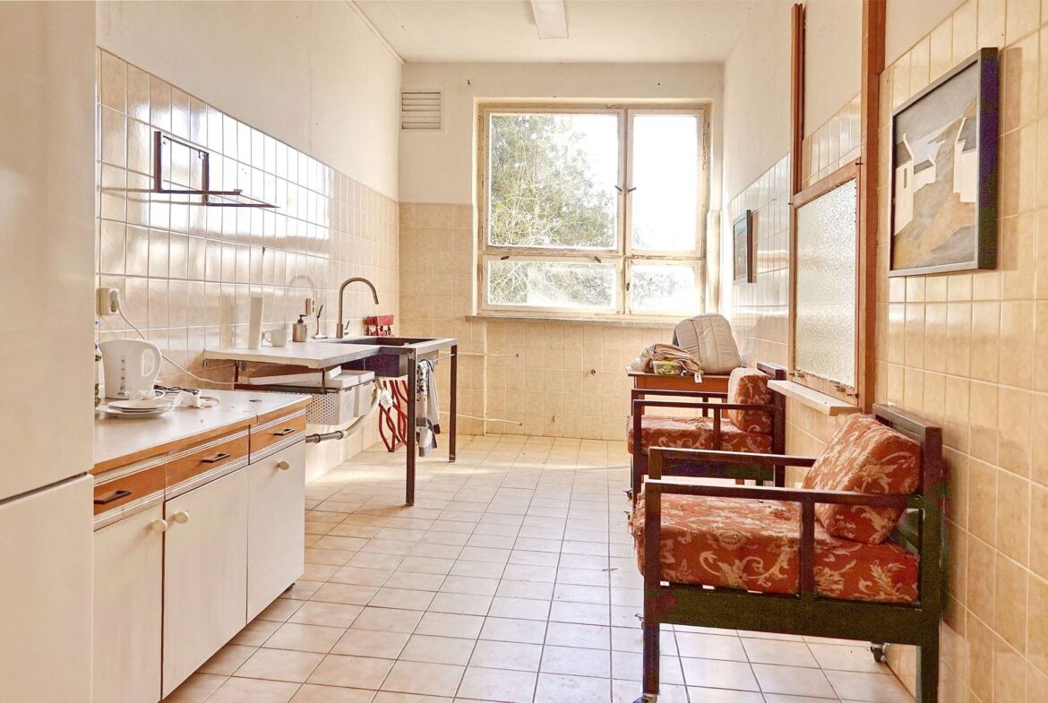 Sunlit retro kitchen with tiled walls, a sink and kettle on the left, and red patterned seating by the window.