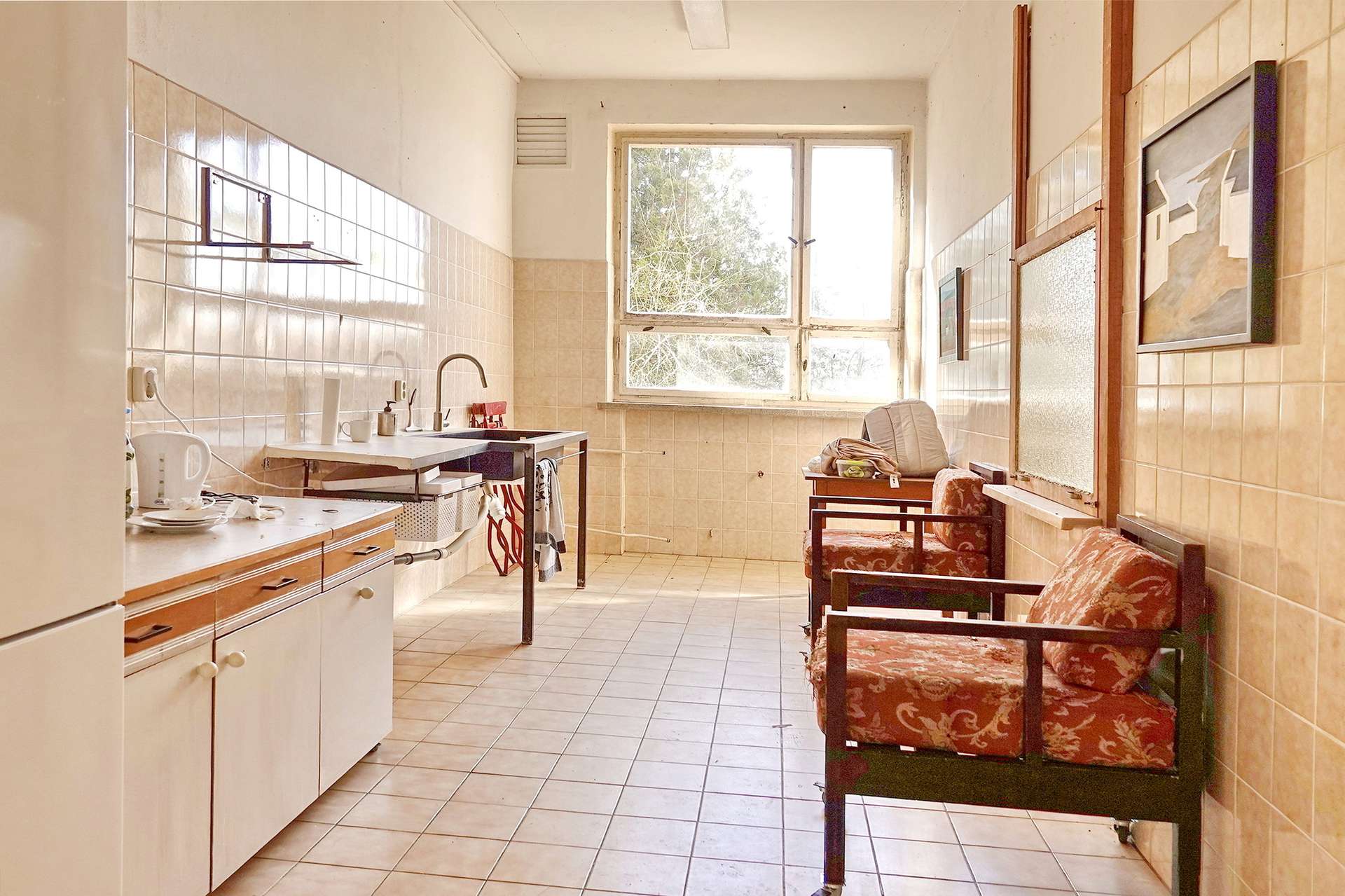 Sunlit retro kitchen with tiled walls, a sink and kettle on the left, and red patterned seating by the window.