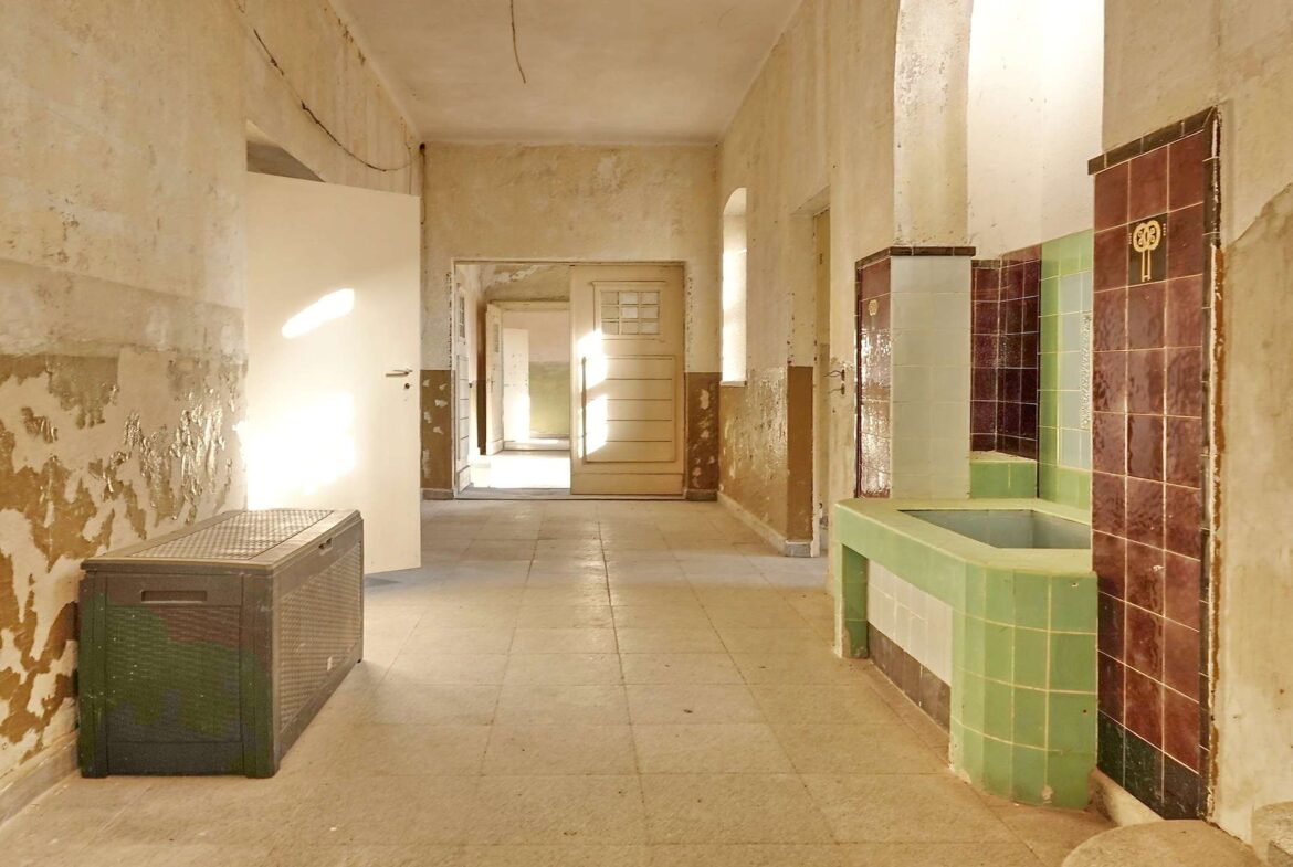 Desolate hallway in an old building with peeling plaster, open doors, and sunlight streaming in; tiled green and brown fixtures along the right wall.