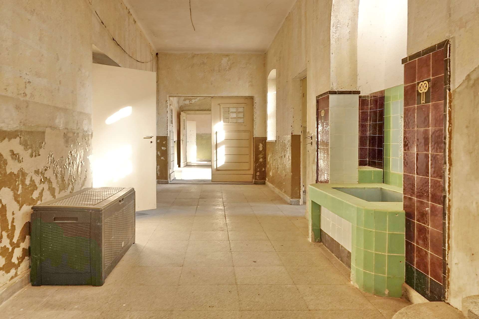 Desolate hallway in an old building with peeling plaster, open doors, and sunlight streaming in; tiled green and brown fixtures along the right wall.
