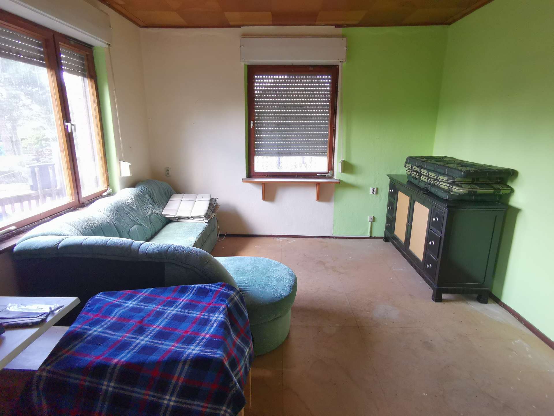 Living room with a blue plaid sofa against a light wall and a green accent wall, window blinds, and a dark cabinet with cushions on top.